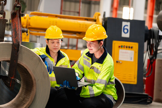Two industrial workers in safety helmets inspecting lifting hook and chain system in factory, checking overhead crane equipment for maintenance and workplace safety compliance.
