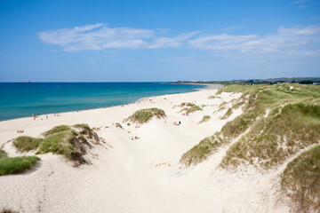 Dunes stretch out at Borestranda beach in Kleppe, Norway, under a clear sky. People enjoy the beach while grass rests on the sandy hills near the coastline.