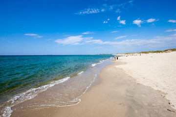 Fototapeta premium People walk along Borestranda beach in Kleppe, Norway. They enjoy the warm sun and the gentle waves. The sandy shore stretches out under a bright blue sky with clouds.