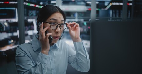 Female Asian Trader Speaks on Smartphone While Executing Stock Market Operations on Computer. Monitoring Live Charts and Price Movements, Woman Reports on Current Market Trends and Fluctuations.