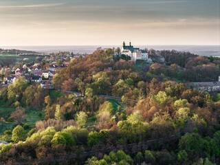 defaultPanorama Góry Świętej Anny o zachodzie słońca. Widok na bazylikę i klasztor na wzgórzu w otoczeniu wiosennej zieleni i kwitnących drzew. Kultowe miejsce kultu i turystyki na Opolszczyźnie. © Henryk Niestrój