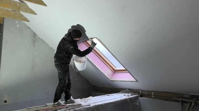 A worker is at an attic in a house applying finishing touches around a window. The light from the window highlights the ongoing renovations and construction work.