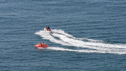 Obraz premium A popular seaside activity is water tubing. A jet ski pulls an inflatable structure with passengers wearing life jackets at high speed.