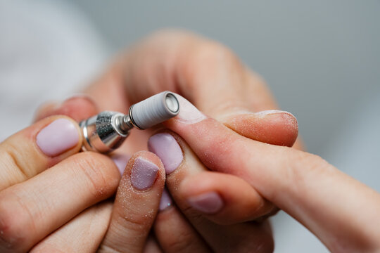 A close-up view of a professional nail technician using an electric milling cutter to shape and file a client's fingernails during a manicure treatment in a beauty salon.