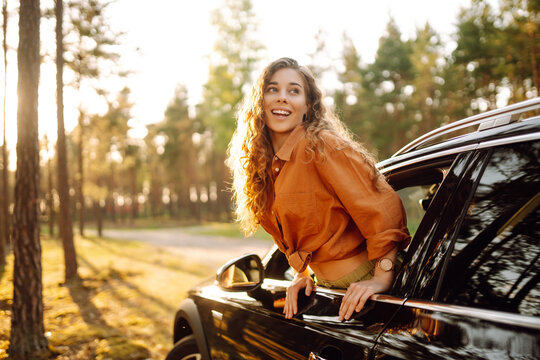 A smiling woman leans out of a car window and enjoys the sunset. A beautiful woman on a car adventure feels free in the sun's rays. The concept of freedom, adventure, and the car.