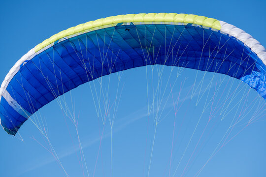 Close-up of a paraglider canopy fully inflated against a clear blue sky. Blue, white and yellow wing with suspension lines, paragliding equipment, air sport and freedom concept.