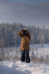 Photographer standing in snowy field taking pictures in winter landscape. Cold weather outdoor activity with forest background and natural daylight.