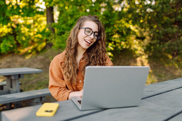 A female freelancer works on a laptop at a wooden table in a sunny park. A young woman sits outdoors with a laptop. Concepts for remote work and education.