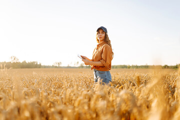 A young farmer with a digital tablet works in a golden wheat field on a sunny day. Female agronomist checks quality of crop in field under sunset. Concept of agriculture, a bountiful harvest. © maxbelchenko