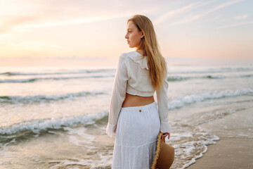 A young woman strolls along the beach at sunrise. A cheerful woman with a hat enjoys the seascape outdoors. Concepts of nature, relaxation, and weekends.