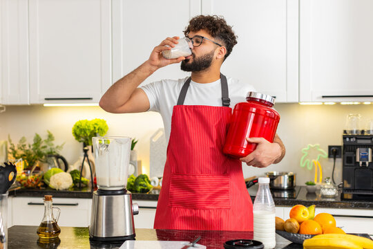 Arabian man pours milk whey protein smoothie with banana and apple into a glass after workout in kitchen at home. Young guy drinks shake smiling enjoying healthy sports nutrition in apartment indoors