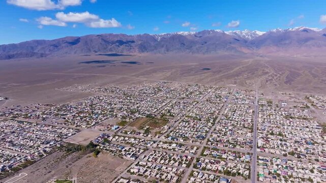 Aerial view reveals expansive residential settlement at foot of snow-capped mountains. Peaceful semi-arid landscape features houses, roads, and trees.