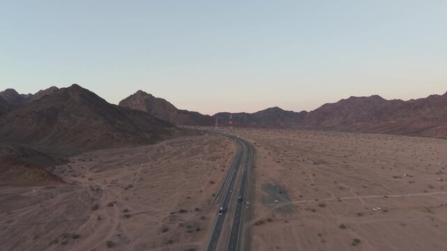 Aerial drone footage of a multilane highway winding through a vast desert valley and rugged mountain range of Wadi Al-Jinn. Vehicles travel along the asphalt road during a serene twilight landscape.