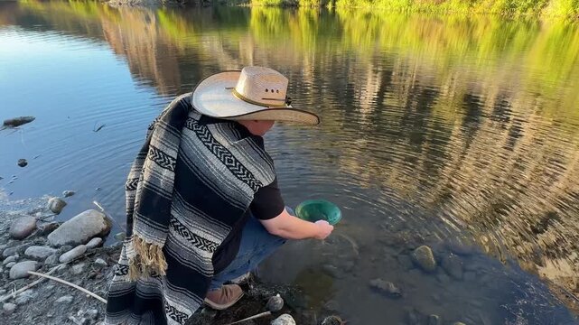 Millennial man (early 40s) wearing a poncho and cowboy hat, panning for gold in Arizona in river
