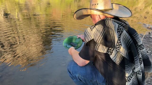 Millennial man (early 40s) wearing a poncho and cowboy hat, panning for gold in Arizona in river