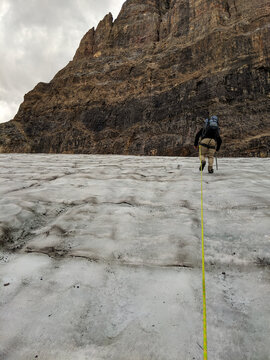 A man wearing a backpack and tethered to a rope, climbs a glacier in the rocky mountains with an icepick during summer.