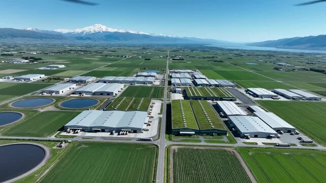 Expansive Aerial View of Modern Warehouses and Green Fields with Mountains in the Background. Perfect for Logistics and Sustainable Business Concepts.