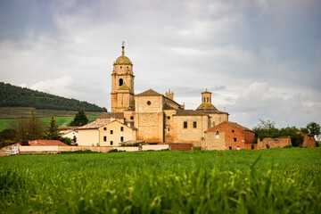 Fototapeta premium Historic stone church and traditional village buildings in rural Spain along the Camino de Santiago, scenic landscape with lush green fields under a cloudy sky in the Spanish countryside.