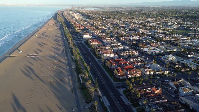 Coastal corridor along PCH parallels wide Huntington City Beach shoreline north of pier district near 15th St. Long palm shadows, clean sand bands, dense roof textures coastal urban divide, CA