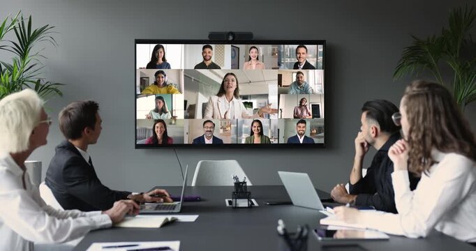 Group of businesspeople gather at conference table collaborating with multiple participants from diverse locations displayed on TV screen, take part in corporate discussion. Business virtual meeting