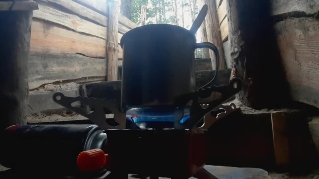 Close-up of a metal mug heating on a portable gas burner inside a wooden field shelter. Survival conditions, outdoor cooking and temporary camp life concept.