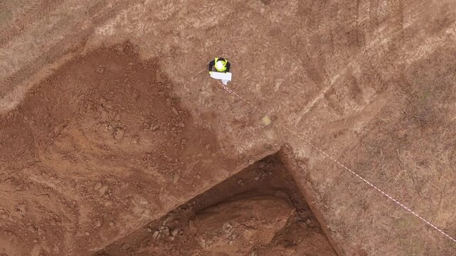 Professional construction supervisor wearing safety helmet and reflective vest reviewing technical documentation next to large excavation pit at building site. Engineering inspection, project planning