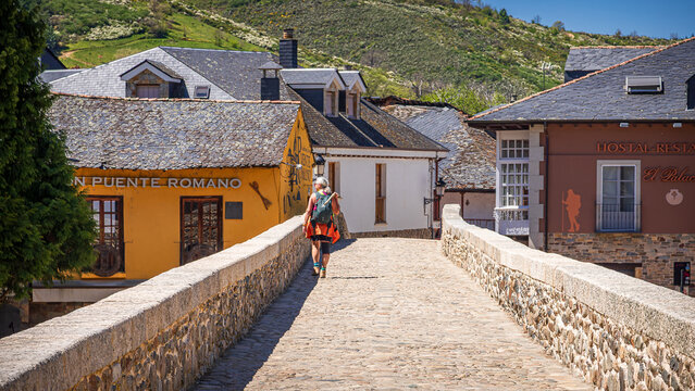 A pilgrim crosses the historic medieval stone bridge leading into the village of Molinaseca, featuring traditional slate-roofed houses and a yellow hostal building, Camino de Santiago, Spain