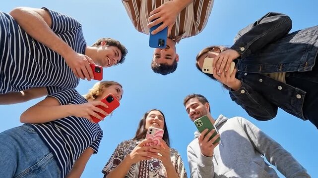 Low angle shot of diverse young friends looking at smartphones in a circle against a bright blue sky, representing digital connection.
