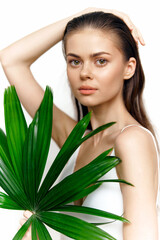Naklejka premium Young woman with green palm leaf, natural beauty, light makeup, emotionless expression, bright background, summer vibes, minimalistic style.