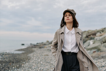 Young woman in a textured coat and hat walks along a rocky beach on a cloudy day. She looks...
