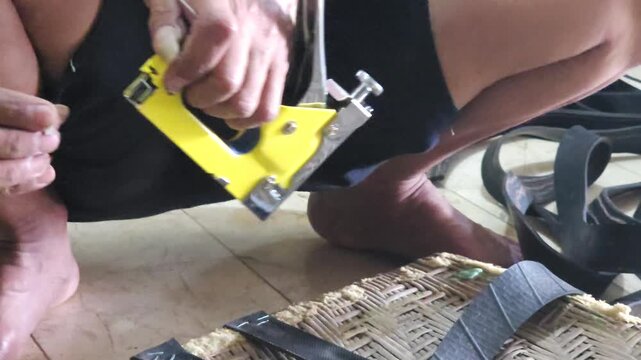 Close up of craftsman hands using yellow staple gun to attach rubber straps to woven chair frame during furniture repair and restoration