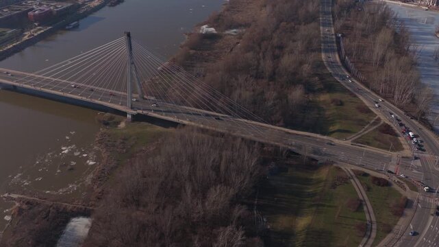 Aerial view shows Swietokrzyski Bridge in Warsaw over the Vistula as traffic moves, ice patches drift, trees are leafless, and long shadows suggest late winter.
