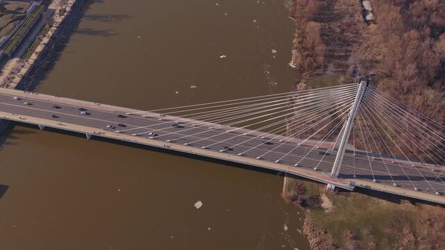Aerial shot shows Swietokrzyski Bridge in Warsaw, single inclined pylon and fan cables, cars and pedestrians in motion, icy river, leafless trees, warm low light.