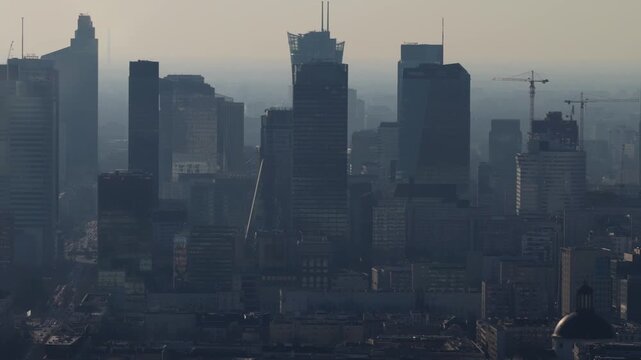 Aerial pan over central Warsaw shows Zlota 44, Warsaw Spire forms, cranes, and a domed landmark. Traffic moves along an avenue in hazy light with layered depth.
