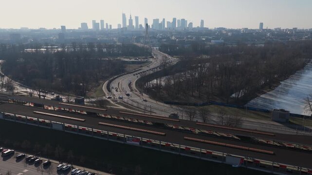 Aerial drone view shows Vistula River, curving multi lane road, A shaped Swietokrzyski Bridge pylon, Varso Tower, Palace of Culture, light traffic, hazy Warsaw skyline.