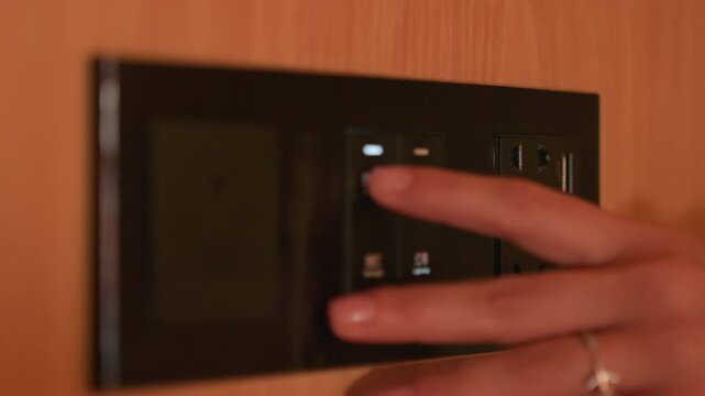 Hotel room control panel closeup, black power panel mounted on wood wall. Hand with ring touches illuminated touch buttons, USB ports and universal outlet visible. Warm ambient lighting creates cozy