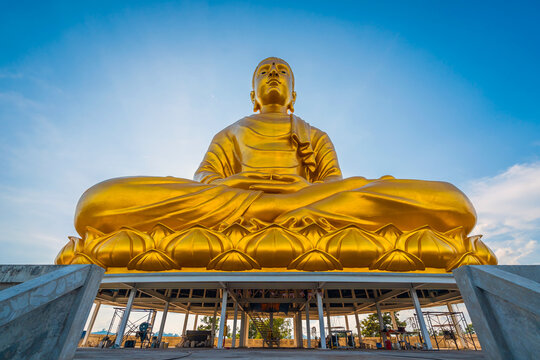 Majestic giant golden Phra Phuttha Lokanat Satsada Buddha statue at Wat Wang Wiwekaram in Sangkhlaburi, Kanchanaburi seated on lotus base beneath radiant blue sky
