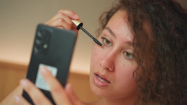 Woman prepares makeup. Woman in hotel room using smartphone as mirror for makeup preparation. Female individual in accommodations utilizing handheld device as mirror for cosmetic application