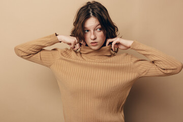 Young woman with short brown hair wearing beige knitted sweater pulls collar up. Neutral background...