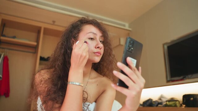 Hotel room guest applies makeup using phone. Warm bedside lighting, wooden wardrobe, wallmounted TV, towelwrapped hair, mirror selfie angle, blending concealer and powder, relaxed evening preparation,