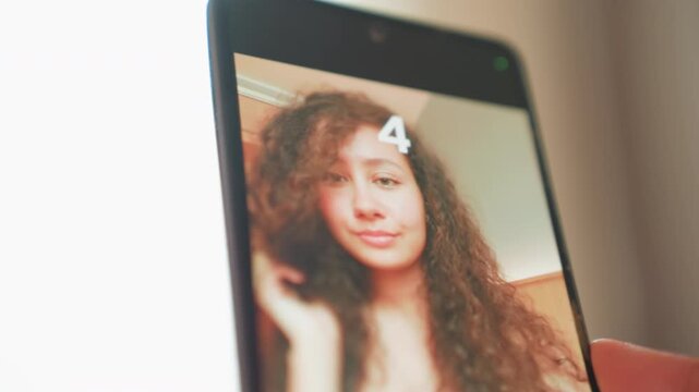 Smartphone selfie showing young woman adjusting hair using timer to capture intimate portrait curly hair, soft warm lighting, indoor bedroom vibe, subtle smile, experimenting with angles