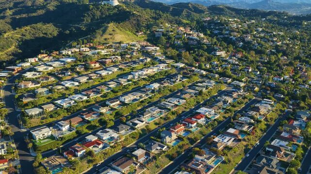 Drone aerial of Hollywood Hills neighborhood with Griffith Observatory visible on hilltop surrounded by green slopes and distant mountains in bright daylight.
