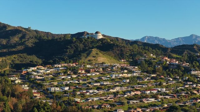 Aerial view of Griffith Observatory rising above green Hollywood Hills with residential neighborhood in foreground and mountain backdrop under clear blue sky.
