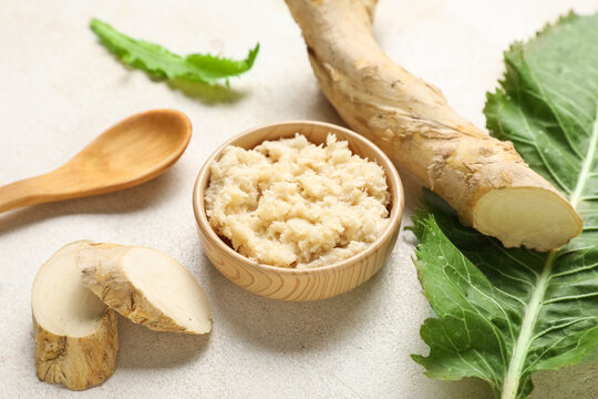 Bowl of horseradish sauce with root, slices, leaf and spoon on light background