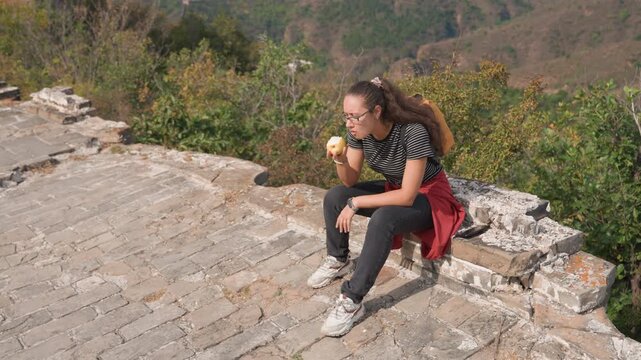 Young woman snacking on ancient walkway, red jacket tied at waist, striped tee, apple in hand, panoramic mountain background, sunlit bricks, passing tourists, playful travel vibe, short rest