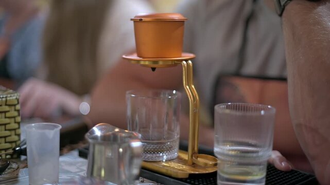Vietnamese Phin Brewing On Table Beside Glass Tray, Golden Filter Perched Above Glass, Blurred Crowd Hands In Background, Casual Street Cafe Vibe, Slow Drip Forming Concentrated Coffee, Warm