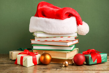 Stack of books with Santa hat, Christmas balls and gift boxes on wooden table against green...