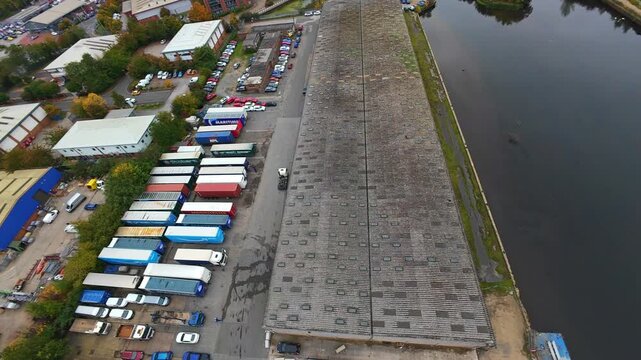 Aerial Pan Over Busy Distribution Yard, Concrete Loading Bays Lined With Lorries, Active Vehicle Movement, Forklift Shifting Pallets, Drone Capturing Shift Change, Riverside Industrial Basin, Autumn