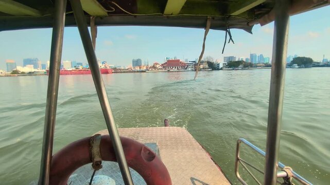 Boat crossing wide Chao Phraya river with Bangkok skyline and Wat Khlong Toei Nok Pier in distance, rearview medium backdrop