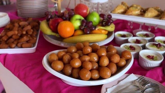 Close up view of a traditional dessert buffet featuring gulab jamun, assorted fresh fruits, syrup pastries and creamy pudding cups arranged on a vibrant indoor catering table.